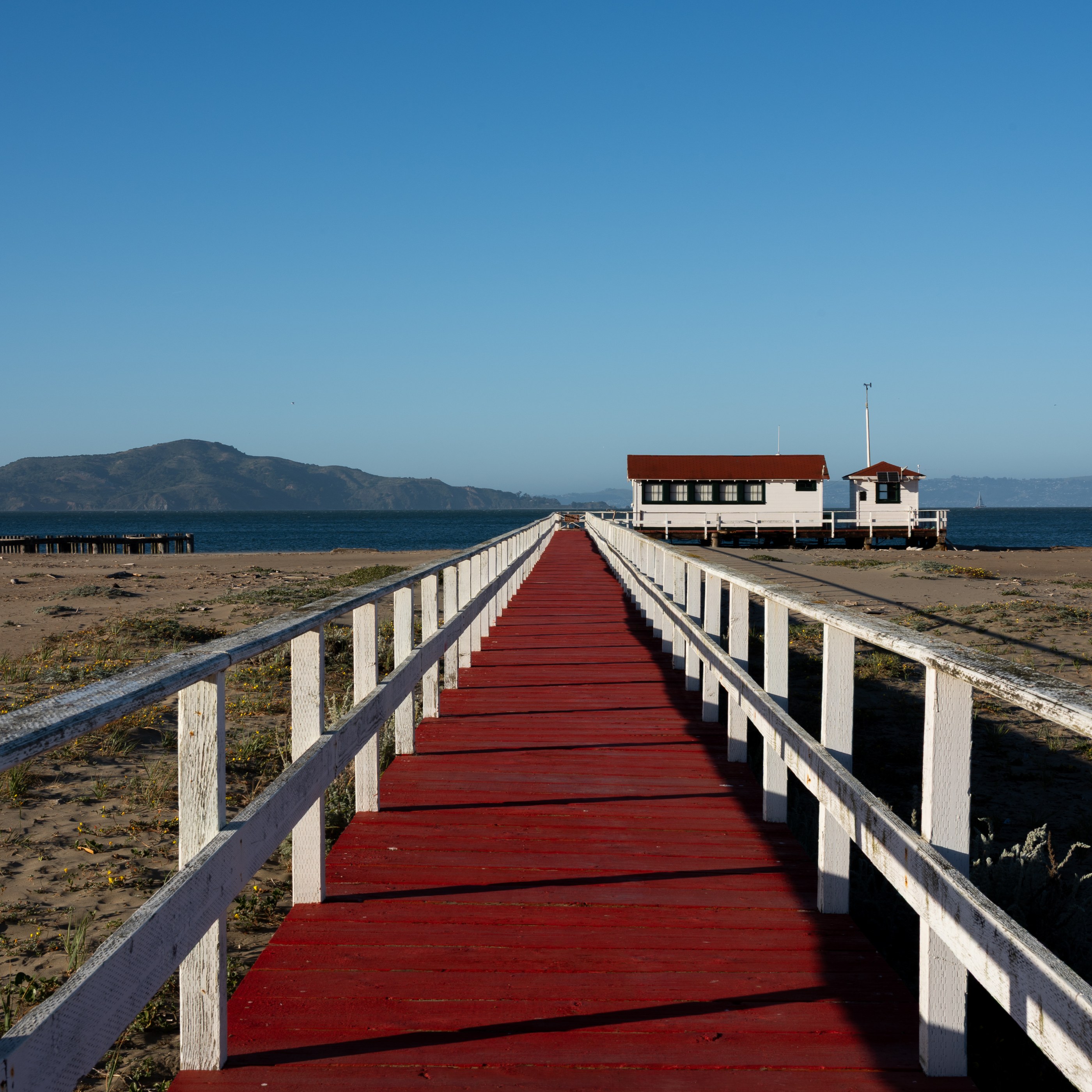 A red wooden walkway extends toward a white building with dark roofing and a small tower, situated near a body of water under a clear blue sky. White railings line both sides of the path. Dry, sandy ground flanks the walkway, and distant