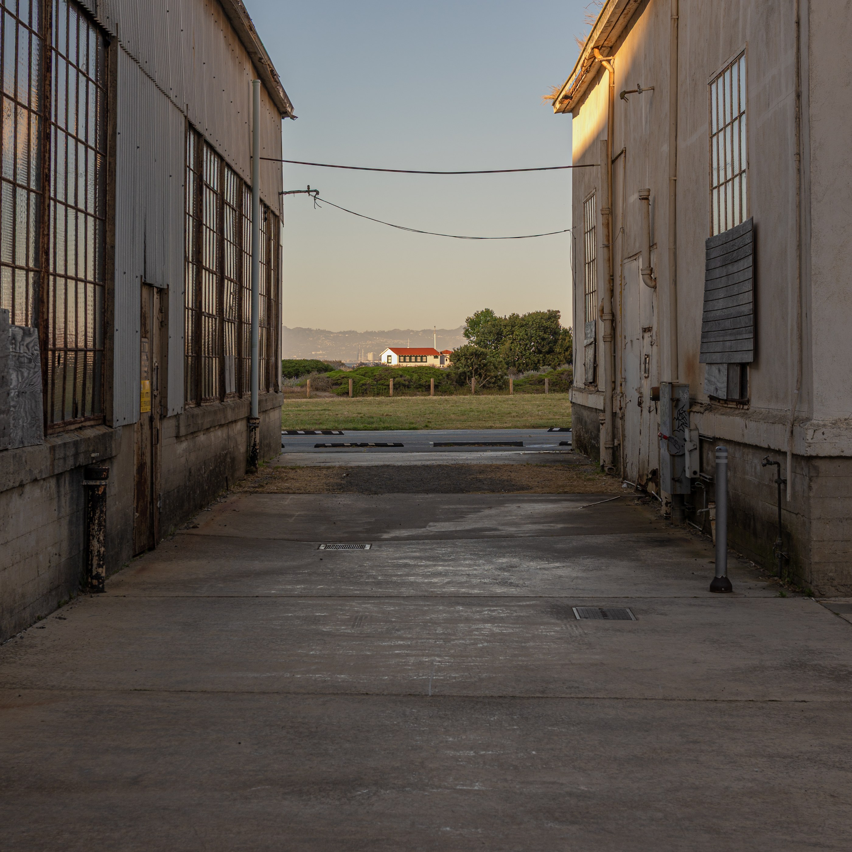 A paved alleyway between industrial buildings leads toward a distant road and buildings, with overhead wires overhead.