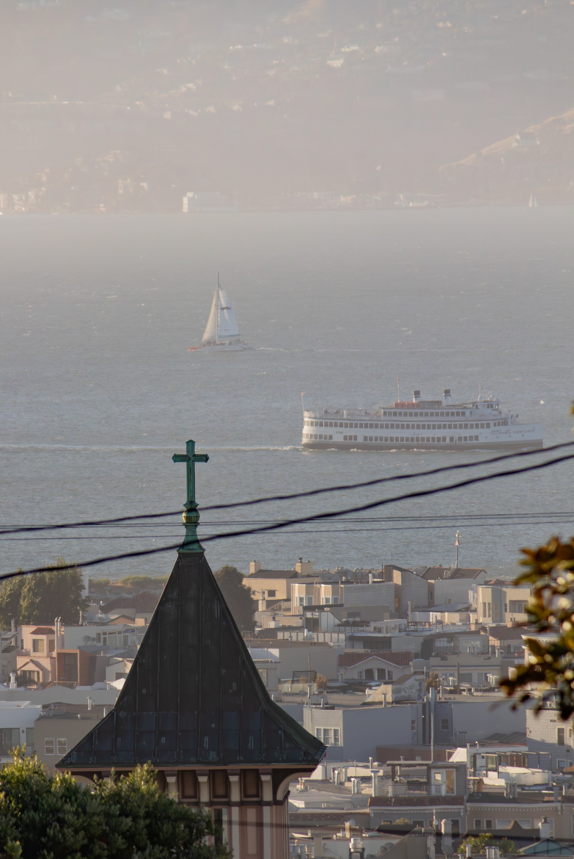 A high-angle view shows a cityscape with a prominent church steeple topped with a dark green cross. Across the water, a large white passenger ship is visible. In the distance, hazy hills rise above the horizon. Power lines cross the