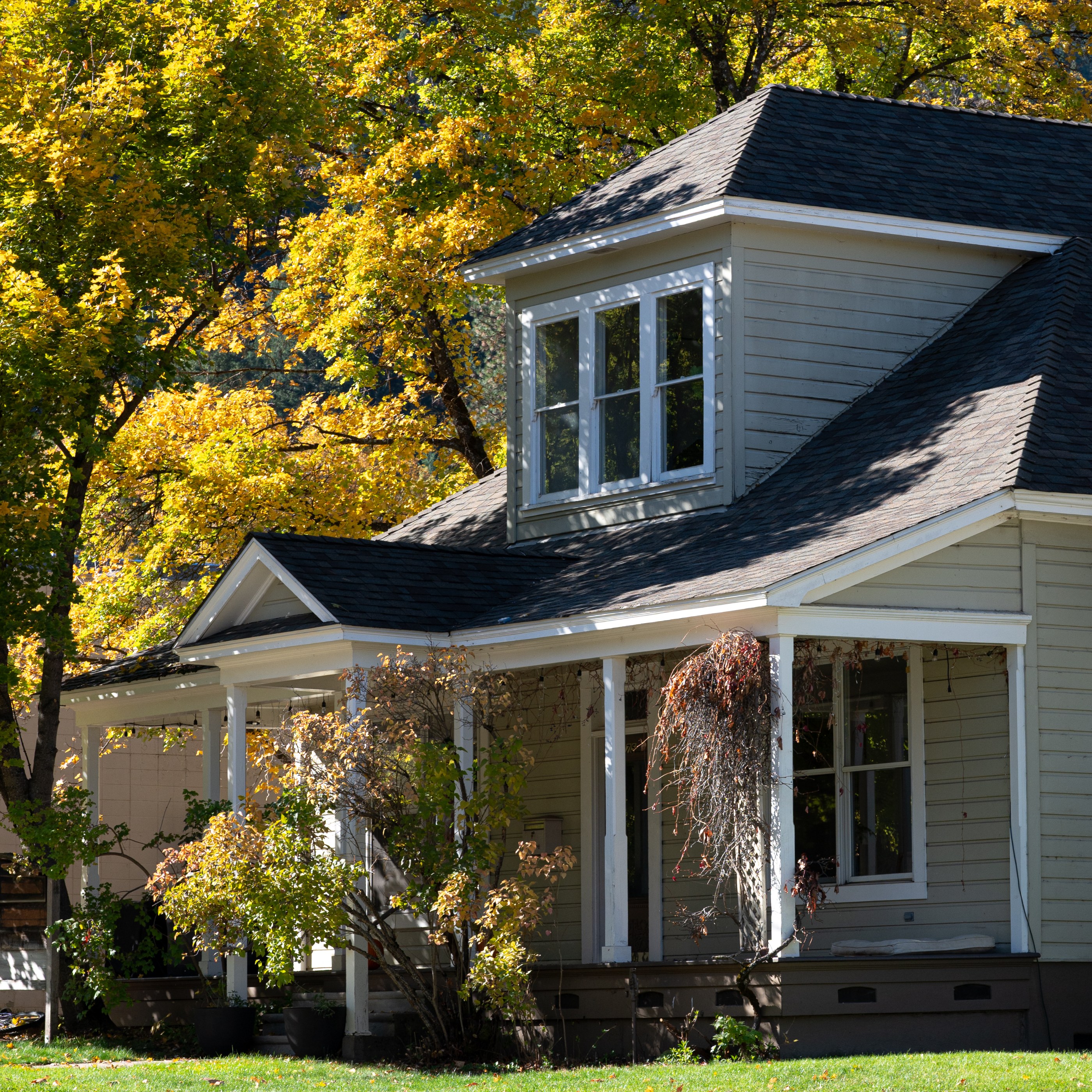A light gray two-story house with a dark gray roof and white trim is surrounded by trees with yellow autumn leaves. A covered porch with white columns and railings extends along the front.
